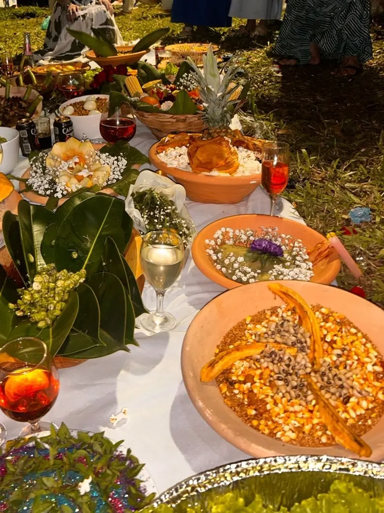 Mesa de oferendas sobre toalha branca, com alguidares de milho e feijão, folhas, flores brancas, frutas e taças, em uma celebração ao ar livre