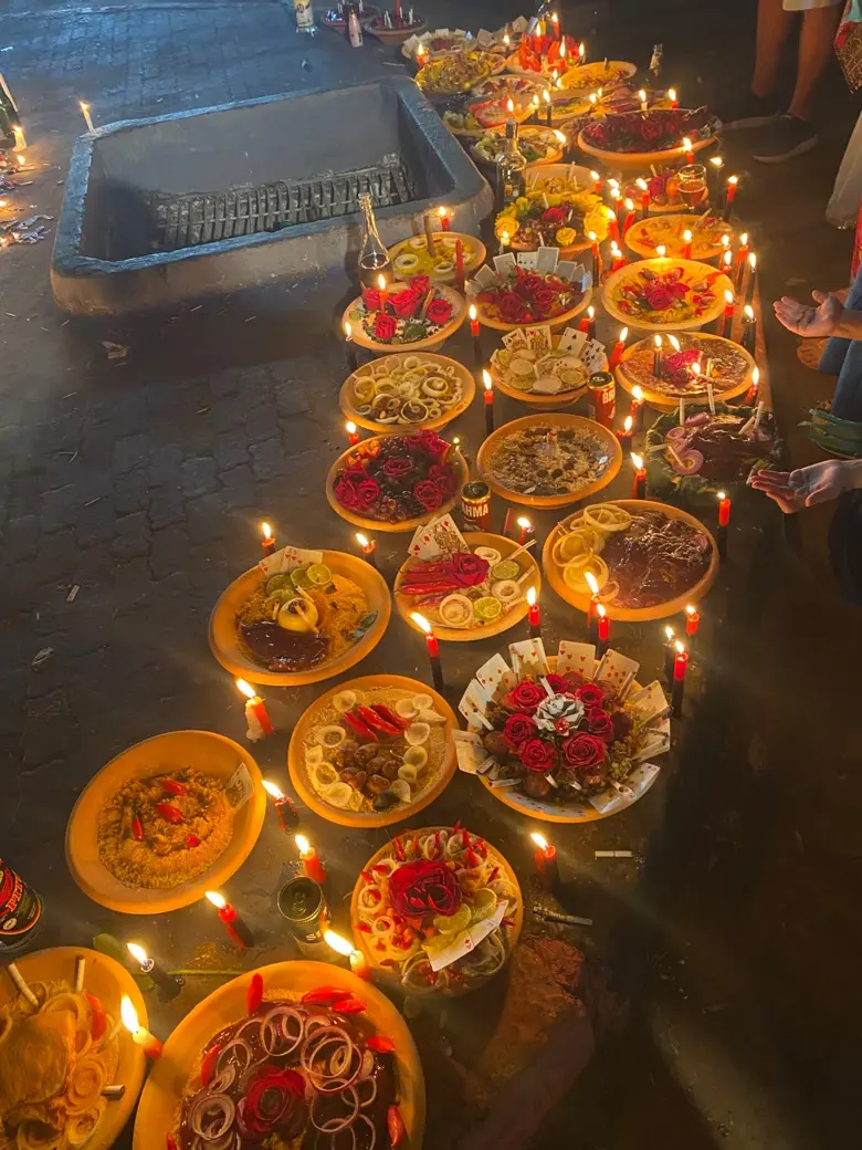 Fileira de alguidares de barro com rosas vermelhas, cebolas, pimentas e cartas de baralho, rodeados de velas vermelhas e pretas acesas