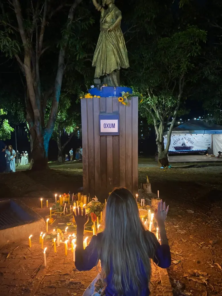 Veronica de costas, com as mãos erguidas, diante da estátua dourada de Oxum cercada de velas amarelas e flores, em uma festa das águas à noite
