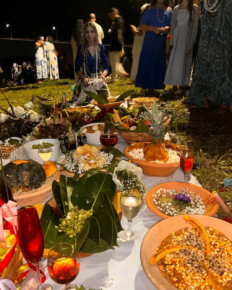 Veronica, com guias no pescoço, sentada atrás de uma mesa de oferendas com frutas, flores brancas, alguidares e taças, em uma celebração ao ar livre à noite